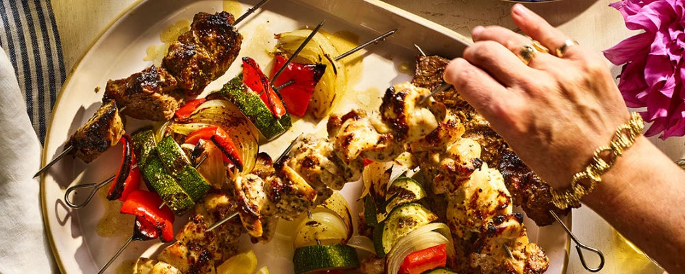 Platter of grilled skewers with vegetables and a hand reaching for one, on a table with a striped cloth.