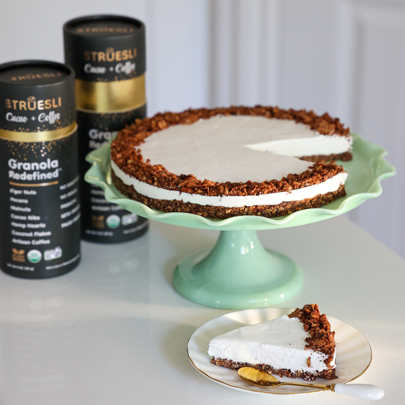 Vegan granola cheesecake on a cake pedestal with containers of Struesli Cacao + Coffee