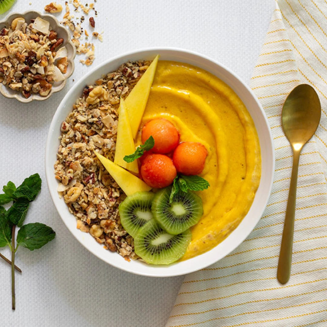 Mango smoothie bowl with granola, fruit, and a spoon on a white surface.