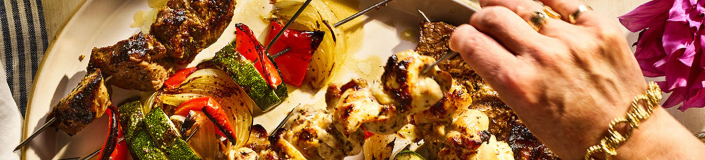Person holding a skewer of grilled vegetables and meat on a plate with a pink flower in the background.